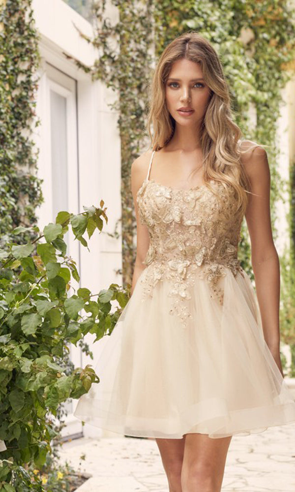 A woman with long wavy hair wears the Short A-Line Butterfly Homecoming Dress 902, featuring beige floral embellishments and spaghetti straps, standing outdoors near greenery and a white building.